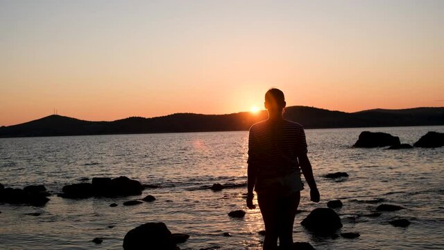 a warm summer evening sunset on the shore of the mora through the strait, the sun almost went down behind the mountains, there are many stones on the near shore and the girl is standing with her back