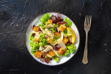 Chicken Caesar salad, shot from the top on a black background. Romaine leaves, crispy croutons, chicken meat, and the classic dressing