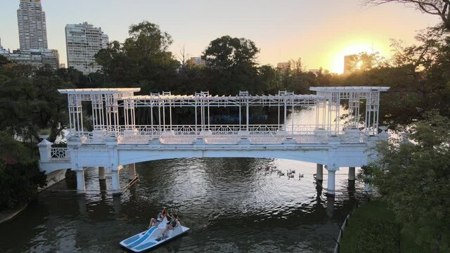 Static shot of tourists paddling paddleboat through bridge followed by a flock of waterfowls in tranquil afternoon with big bright sun setting at the background at rose garden Parque Tres de Febrero.