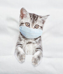 Kitten wearing medical protective mask lying under white warm blanket on a bed at home. Top down...