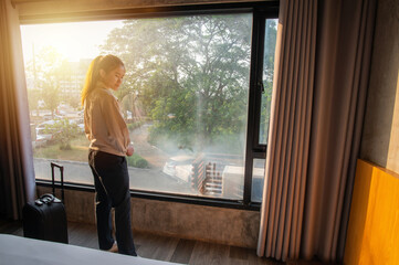 Portrait of tourist woman standing nearly window with her luggage in bedroom after check-in in hotel. Conceptual of woman lifestyle while traveling and vacation.