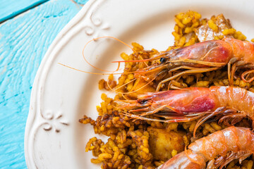 Detail of a portion of typical Spanish paella with rice and seafood, with three prawns on top, in an old white plate on a blue background