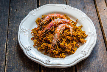 A portion of typical Spanish paella with rice and seafood, with three prawns on top, in an old white plate on a wooden board