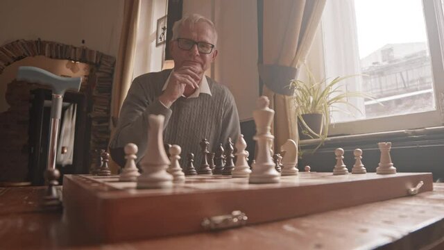 Slowmo POV shot of thoughtful senior man playing chess game on board on wooden table in nursing home