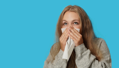 Young blond woman suffering from allergy on light blue background. Symptoms of cold or allergy. Woman sneezes in white tissue. - obrazy, fototapety, plakaty