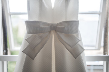 Beautiful pretty big bow on front of brides bright elegant white wedding dress hanging up in the window of the bridal suite on the morning of the ceremony.