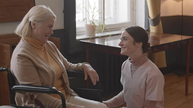 Slowmo shot of young caring nurse and smiling senior woman in wheelchair having conversation in nursing home