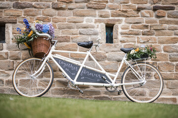 Wedding vintage old retro tandem bike with just married sign and fresh flowers in woven basket. Beautiful cream white bicycle at marriage venue with roses and petals leant against stone wall barn.