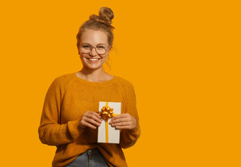 Smiling beautiful blonde young woman wearing eyeglasses holding a gift box isolated over yellow background. Happy birthday, Christmas, Valentine's Day, vision concept. Place for text.
