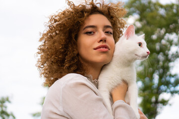 A beautiful teenage girl with curly hair holds a white cat close to her face. She is enjoying time with her pet.