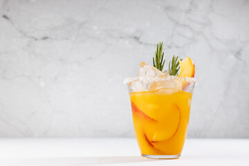Refreshing juicy peach cocktail with ice, rosemary, sugar rim, fruit slices in misted glass on white wood table, concrete wall.
