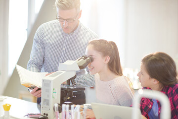 Male teacher helping girl students using microscope, conducting scientific experiment in laboratory classroom
