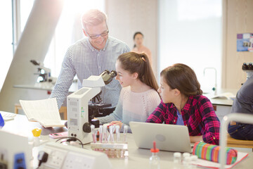 Fototapeta premium Male teacher helping girl students using microscope, conducting scientific experiment in laboratory classroom