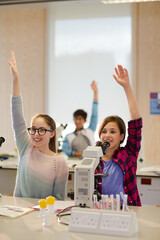 Eager students raising arms behind microscopes in laboratory classroom