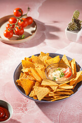 A blue bowl with cheese tortilla chips and a chilli cheese dip on a pink marble surface, cherry tomatoes, ketchup in hard morning light