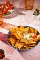 A blue bowl with cheese tortilla chips and a chilli cheese dip on a pink marble surface, cherry tomatoes, ketchup in hard morning light