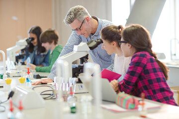 Male teacher helping girl students at microscope in laboratory classroom