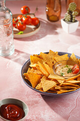 A blue bowl with cheese tortilla chips and a chilli cheese dip on a pink marble surface, cherry tomatoes, ketchup in hard morning light