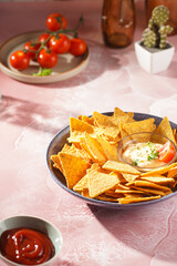 A blue bowl with cheese tortilla chips and a chilli cheese dip on a pink marble surface, cherry tomatoes, ketchup in hard morning light