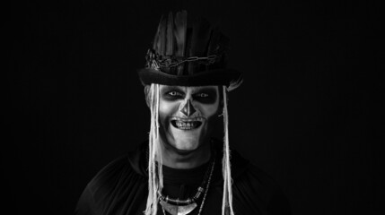 Creepy man with skeleton makeup in top-hat with feathers. Guy making faces, trying to scare, toothy smile. Voodoo rituals. Baron Saturday. Halloween thematic party. Black and white image