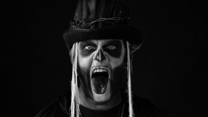 Frightening man in skeleton Halloween cosplay costume. Fashion art design. Guy in creepy skull makeup making faces, showing tongue, trying to scare. Day of The Dead. Black background. Black and white