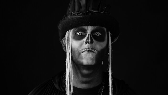 Sinister man with Halloween skeleton makeup in costume with top-hat turning his head and looking at camera trying to scare. Horror theme. Day of The Dead. Isolated on black background. Black and white