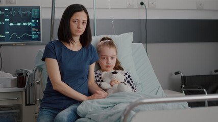 Portrait of worried mother sittting beside hospitalized little child looking into camera during medical examination in hospital ward. Sick girl resting in bed recovering after recovery surgery