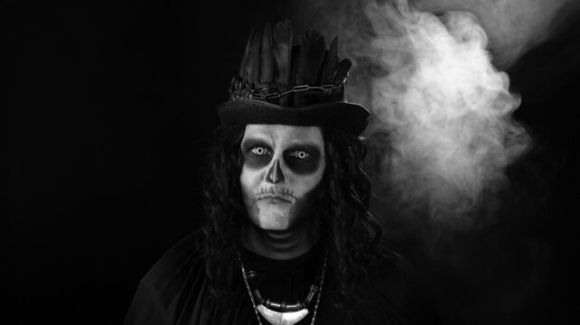 Creepy long hair man with skeleton makeup in top-hat with feathers. Guy making faces, trying to scare, toothy smile. Voodoo rituals. Baron Saturday. Halloween party. Black background. Black and white