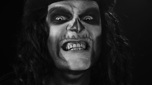 Close-up of frightening man face in skeleton Halloween cosplay. Crazy guy in creepy skull makeup looking at camera, showing dirty black teeth, trying to scare smiling. Dark background. Black and white