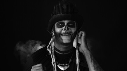 Scary guy in thematic carnival costume of Halloween skeleton wearing earphones listening music dancing celebrating against dark background. Sinister man with professional skull makeup. Black and white