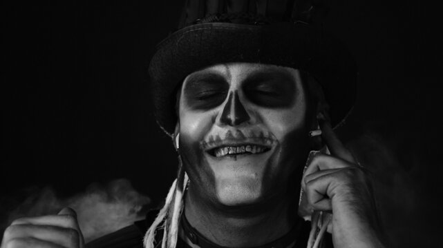 Sinister man with horrible Halloween skeleton makeup with top-hat puts on headphones and starts dancing, celebrating. Horror theme. Day of The Dead. Isolated on dark black background. Black and white