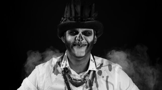 Creepy man with skeleton makeup in top-hat. Guy looking at camera, trying to scare. Voodoo rituals. Baron Saturday. Halloween thematic party. Black background. Black and white portrait