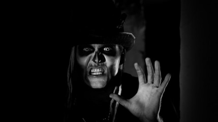 Frightening man with horrible Halloween skeleton makeup in costume with top-hat appears from dark...