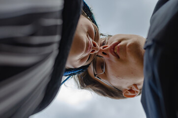 A heterosexual couple in love kissing against the sky. A young man wearing optical glasses. Young woman with braided thin pigtails. View from bottom to top. Selective focus.