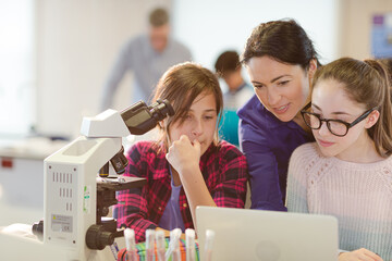 Female teacher and girl students using microscope and laptop, conducting scientific experiment in...