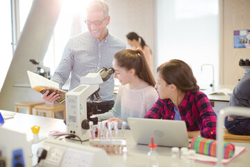 Male teacher helping girl students using microscope, conducting scientific experiment in laboratory classroom