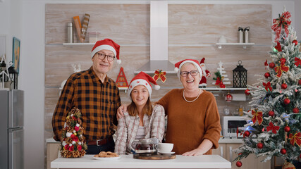 Pov of happy family wearing santa hat greeting remote friends during online videocall conference meeting standing at table in xmas decorated kitchen. Grandparents enjoying christmas holiday