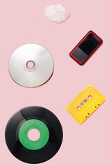 Music mediums over time concept. EP and LP vinyl, cassette tape, CD, mp3 player, cell phone, portable speaker cotton cloud shaped prop flat lay arrangement against pink background.