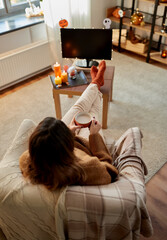 halloween, holidays and leisure concept - young woman watching tv and drinking hot chocolate with her feet on table at cozy home
