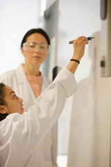 Female science teacher and girl student writing at whiteboard in classroom