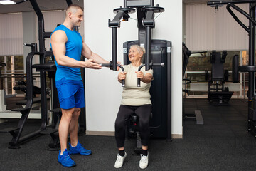 Fototapeta na wymiar handsome young coach man shows senior woman how to properly perform exercises on the simulator in the gym. Happy people smile, enjoy, happy. Active sports healthy lifestyle indoor. Old