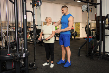 Fototapeta na wymiar young handsome coach man helps a pensioner to do an exercise on the simulator in the gym. Happy people smile. Active healthy sports lifestyle, senior concept.