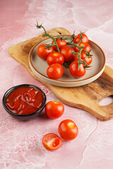 Several red cherry tomatoes with a branch on a wooden board on a pink marble surface and ketchup