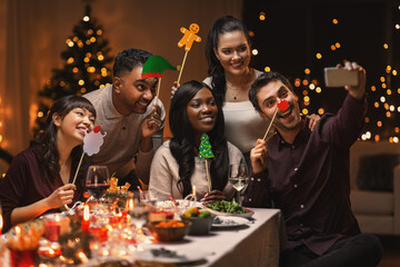 holidays, party and celebration concept - multiethnic group of happy friends having christmas dinner at home and taking selfie with smartphone