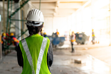 Young Asian female construction engineer wearing a hat and a green safety vest, stands looking at work in the construction zone.