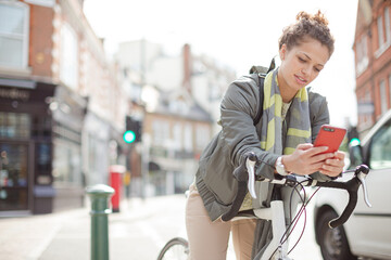 Obraz premium Young woman commuting on bicycle, texting with cell phone on sunny urban street