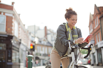 Young woman commuting on bicycle, texting with cell phone on sunny urban street