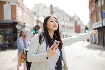 Young businesswoman with cell phone hailing taxi on sunny urban street