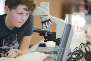 Boy student conducting scientific experiment at microscope and computer in laboratory classroom