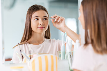 beauty, make up and cosmetics concept - teenage girl with tweezers tweezing her eyebrow and looking to mirror at home bathroom